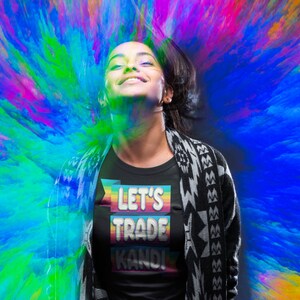 May include: A person wearing a black t-shirt with a rainbow-colored banner that says "Let's Trade Kandi". The person is standing in front of a colorful, abstract background.