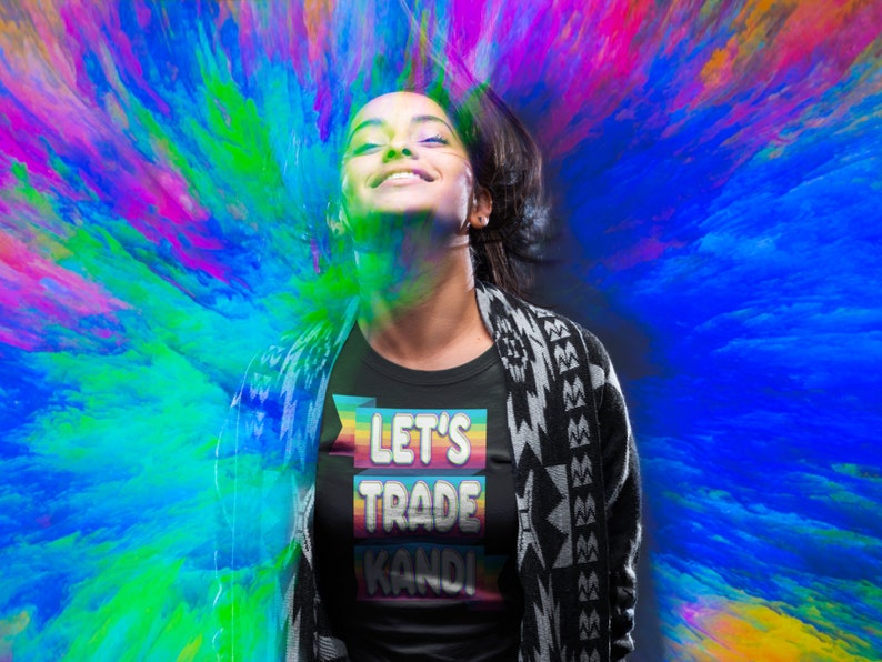 May include: A person wearing a black t-shirt with a rainbow-colored banner that says "Let's Trade Kandi". The person is standing in front of a colorful, abstract background.
