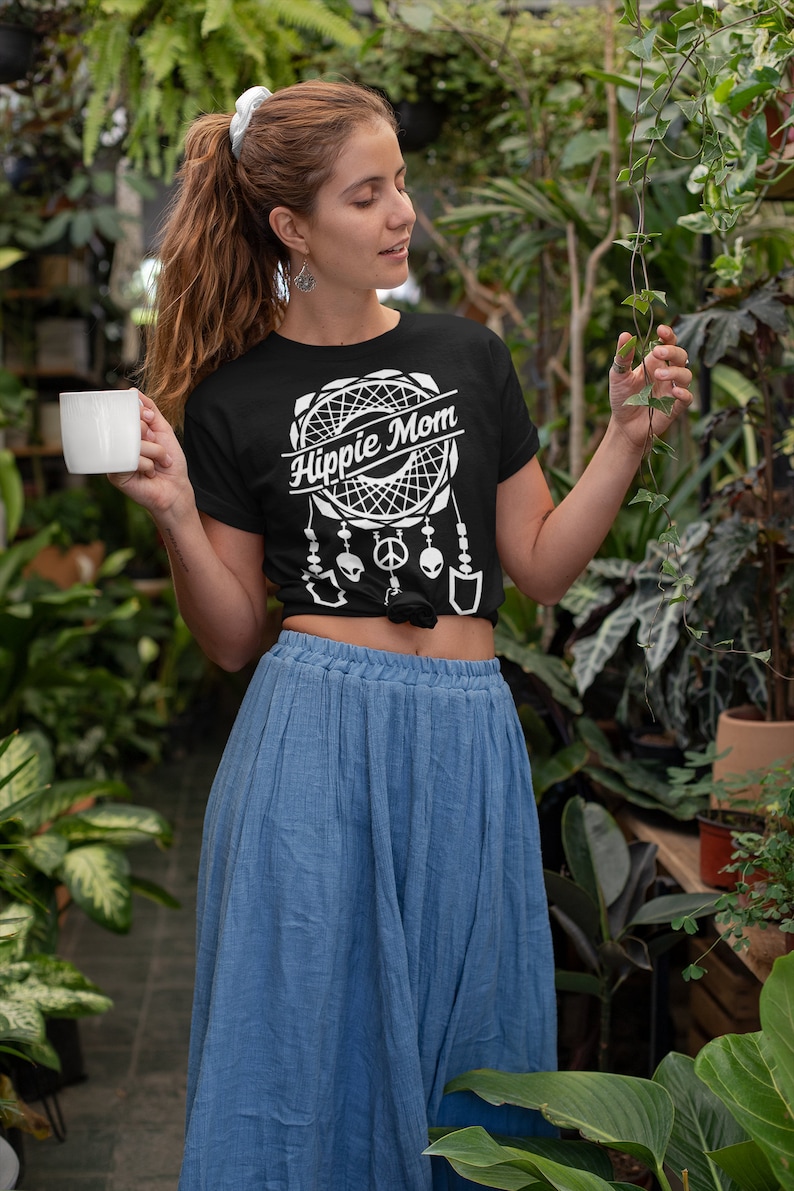 Może przedstawiać: Kobieta w czarnym t-shircie z białym wzorem łapacza sn&oacute;w i napisem "Hippie Mom". Ma na sobie niebieską sp&oacute;dnicę i trzyma biały kubek.