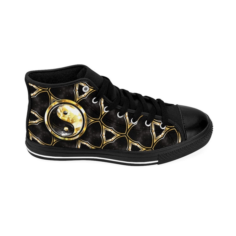 May include: Black high-top sneakers with a gold and black geometric pattern and a gold Yin Yang symbol. The shoes have black laces, a black toe cap, and a white stripe along the sole.