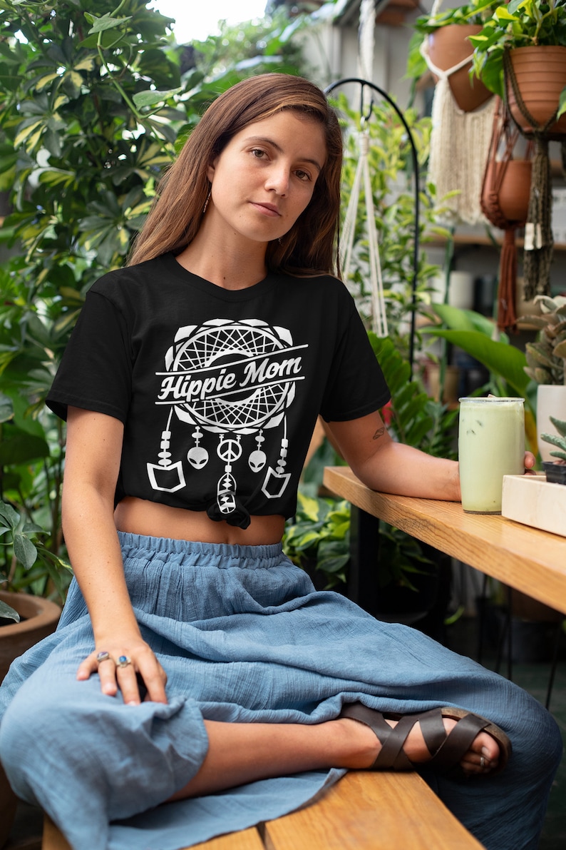 Może przedstawiać: Kobieta w czarnym t-shircie z białym wzorem łapacza sn&oacute;w i napisem "Hippie Mom". Ma na sobie niebieską sp&oacute;dnicę i brązowe sandały.