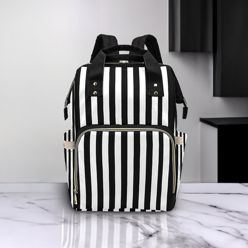 May include: A black and white striped backpack with multiple compartments and gold-colored zippers. The bag has two shoulder straps and a top handle. The design features vertical stripes and a front pocket.