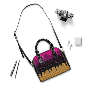 Puede incluir: Un bolso negro con un dise&ntilde;o de cono de helado rosa y negro. El bolso tiene una correa larga y un asa superior.