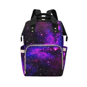 Pode incluir: Uma mochila para fraldas preta e roxa com estampa de gal&aacute;xia, com um bolso frontal com z&iacute;per e al&ccedil;as ajust&aacute;veis.