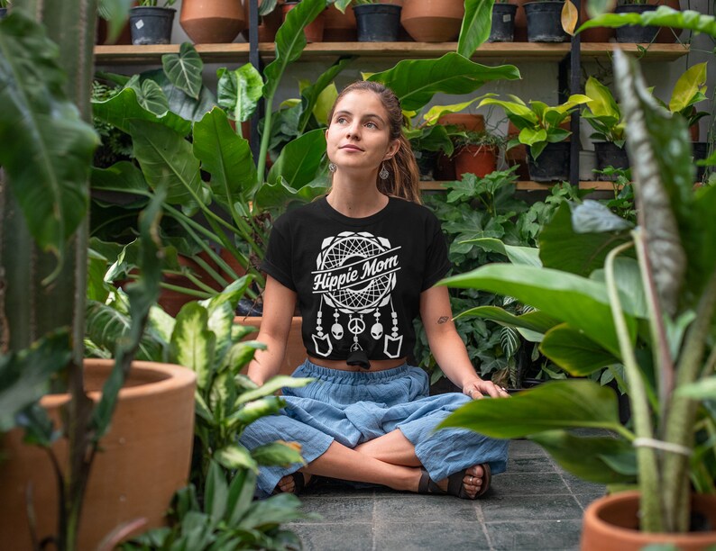 Może przedstawiać: Kobieta w czarnym t-shircie z białym wzorem łapacza sn&oacute;w i napisem "Hippie Mom". Siedzi w pozycji skrzyżnej w bujnym zielonym ogrodzie.