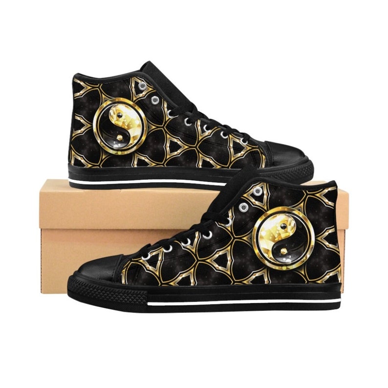 May include: Black high-top sneakers with a gold and black geometric pattern and a gold yin-yang symbol. The shoes have black soles with a white stripe and white laces. The sneakers are displayed on a brown cardboard box.