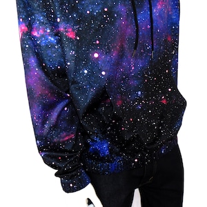 May include: A black hoodie with a galaxy print featuring shades of blue, pink, and white. The hoodie has a drawstring hood and a kangaroo pocket.