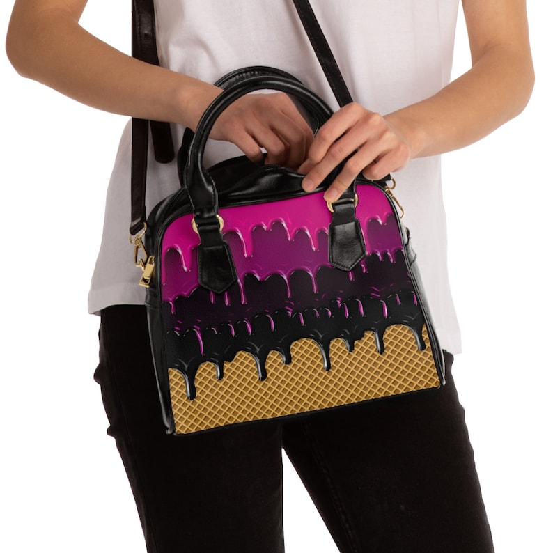 Puede incluir: Un bolso negro con un dise&ntilde;o de cono de helado rosa y negro. El bolso tiene una cremallera dorada y una correa de cadena dorada.