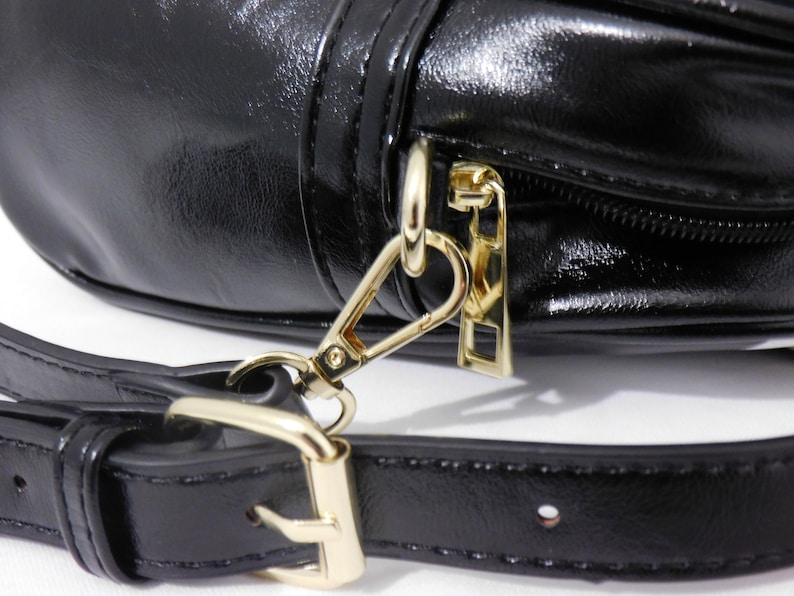 Puede incluir: Primer plano de un bolso de cuero negro con una cremallera dorada y un cierre dorado. El bolso tiene una correa de cuero negro.