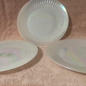 May include: Three iridescent white glass plates. One is round, and two are oval-shaped. The plates have a textured, ridged design around the edges. The plates are on a pinkish-brown surface.