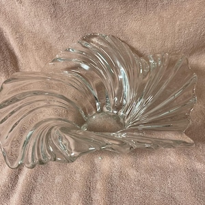 Puede incluir: Un cuenco de cristal transparente con un diseño en forma de abanico giratorio con crestas profundas y radiantes que se extienden desde el centro hasta los bordes ondulados y festoneados. El cristal es liso, creando una forma fluida y dinámica.