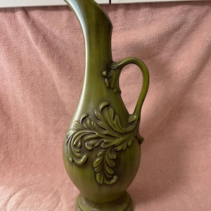 May include: Olive green ceramic pitcher with a curved spout and handle. The pitcher features a raised floral design on the front and sits on a round base. A decorative piece for the home.
