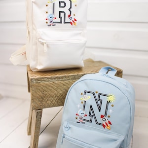 May include: Two small backpacks, one white and one light blue, with embroidered space-themed designs. The white backpack features a large black letter "R" with a rocket, astronaut, and sun. The blue backpack has a black "N" with similar space motifs.