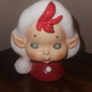 May include: A ceramic Christmas elf figurine with a white head and hair, green eyes, and a red bow. The elf is wearing a red outfit and holding a white ball. The figurine has a vintage, retro style.