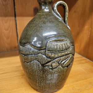 May include: A green ceramic jug with a handle and a carved design of a barrel on the side.