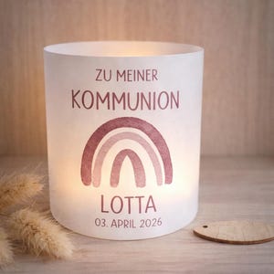 Könnte beinhalten: Ein weißer Kerzenhalter mit dem Text "ZU MEINER KOMMUNION" und dem Namen "LOTTA" in Malve gedruckt. Ein malvenfarbenes Regenbogen-Design befindet sich unter dem Text. Das Datum "03. APRIL 2026" ist unter dem Namen aufgedruckt. Der Kerzenhalter steht auf einer Holzoberfläche.
