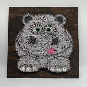 Può includere: Un ippopotamo grigio in arte di filo con occhi verdi e una bocca rosa su uno sfondo di legno marrone scuro.