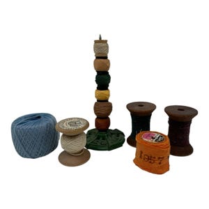 Peut inclure: Une collection d'accessoires de couture comprenant une pelote de fil bleu clair, une bobine en bois avec du fil blanc, un support en métal vert portant six petites bobines de fil coloré, deux bobines en bois et un rouleau de tissu orange.