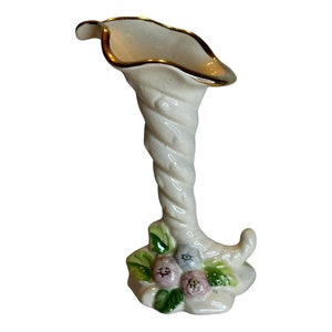 Puede incluir: Un jarrón de cerámica blanca con un borde dorado y un diseño retorcido. La base del jarrón está decorada con flores rosas y azules.