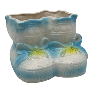 Könnte beinhalten: Keramik-Babyschuh-Pflanzgefäß in Weiß und Hellblau. Der Pflanztopf hat ein strukturiertes Design mit blauen Schleifen und gelben Akzenten. Der obere Rand ist gewellt.