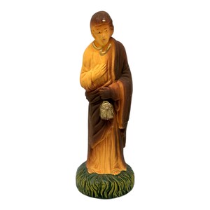 Puede incluir: Una figurita de una persona vestida con una túnica marrón y amarilla, sosteniendo un pequeño objeto dorado. La figurita está de pie sobre una base verde con detalles de hierba.