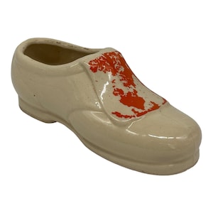 May include: A ceramic shoe planter in a light beige color with an orange design on the side. The shoe is shaped like a classic men's dress shoe.
