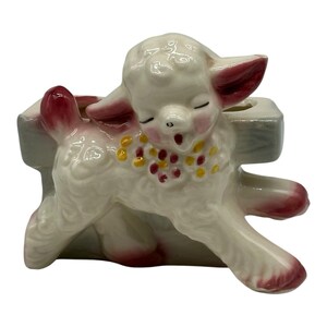 Könnte beinhalten: Ein Keramik-Pflanzgefäß in Form eines Lammes, das vor einem Zaun steht. Das Lamm ist weiß mit rosa Akzenten an Ohren, Schwanz und Hufen und trägt ein gemaltes Halsband aus gelben und roten Blumen um den Hals, die Augen sind geschlossen.