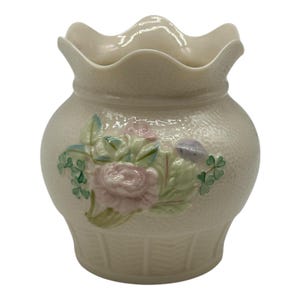 May include: Cream-colored ceramic vase with a textured surface and a ruffled rim. The vase is adorned with a floral design featuring pink roses, green leaves, and purple accents. The base has a basket weave design.