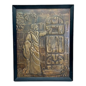 Op de afbeelding: Een ingelijst reliëf van een metalen wandkunstwerk dat een figuur in een gewaad voorstelt die naar een gesloten deur reikt. Het kunstwerk is in tinten bruin en goud.