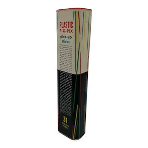 Peut inclure: Une boîte en métal vintage pour un jeu de bâtonnets. La boîte est blanche avec des lettres noires et rouges qui indiquent "Plastic Pix-Pix Pick-up Sticks". La boîte a un fond rayé noir et blanc avec des bâtonnets colorés qui dépassent du haut.