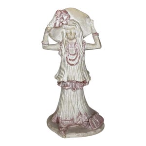 May include: A white ceramic figurine of a woman wearing a white dress with pink accents. She is holding a large, white hat over her head. The figurine is standing on a base with a pink and white design.