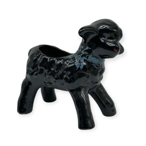 May include: A glossy black ceramic planter shaped like a lamb, featuring a textured, curly wool surface and a smooth, open top. The figure has a small patch of blue-grey paint detail on the neck area and stands on four sturdy, short legs.