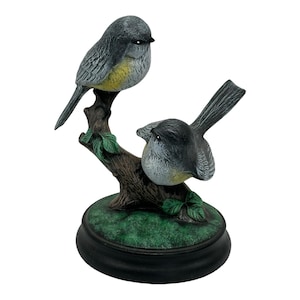 May include: A decorative figurine featuring two small birds with grey and yellow plumage perched on a brown textured tree branch. The branch is set upon a circular green base with a black border, showcasing detailed feathers and green leaves.