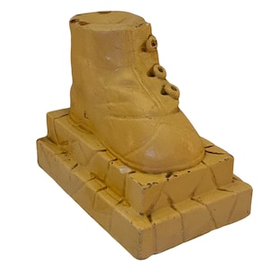 May include: A yellow ceramic boot figurine with a raised base. The boot has a lace-up design with three holes for the laces.