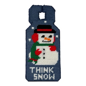 以下が含まれることがあります： 紺色のクロスステッチのクリスマスオーナメント。ドアハンガーの形をしており、黒い帽子、赤いイヤーマフ、緑のマフラー、赤いミトンを身に着けた雪だるまが描かれています。「THINK SNOW」の文字が下部に刺繍されており、背景には雪の結晶があります。