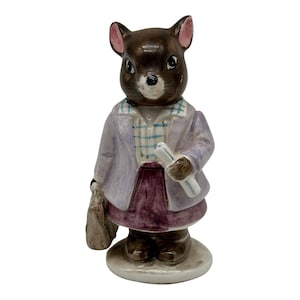 Puede incluir: Figura de cerámica de un ratón vestido con una chaqueta lila claro, camisa a cuadros y falda burdeos. El ratón sostiene un papel enrollado y un bolso. La figura tiene un cuerpo marrón, orejas rosas y está sobre una base redonda.