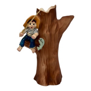 May include: A ceramic vase shaped like a tree trunk with a boy figurine sitting on a branch. The boy is wearing a blue and white striped shirt, blue pants, and a yellow hat. He is holding a fishing rod and there are two fish on the tree trunk.