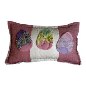 Op de afbeelding: Een roze en wit gestreept kussen met drie patchwork-paaseieren erop genaaid. De eieren zijn paars en groen, paars en wit, en roze en wit.