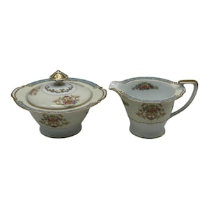 Puede incluir: Azucarero de porcelana color crema con tapa y jarra para crema. Ambas piezas presentan un diseño floral con detalles en rosa, amarillo y azul, y ribete dorado. El azucarero tiene un adorno dorado en la tapa.