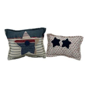May include: Two decorative pillows. One pillow features a star design with red, white, and blue accents. The other pillow has two navy blue star appliqués on a white background with red floral patterns. Both pillows have a rectangular shape.