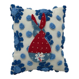 Può includere: Un cuscino decorativo quadrato con sfondo bianco, adornato con dettagli floreali e pompon blu. Un design a coniglietto patchwork a pois rossi e denim blu è centrato sul cuscino, con una piccola applicazione floreale.
