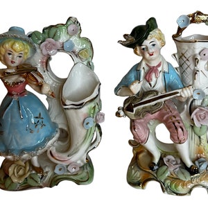 May include: A pair of vintage porcelain figurines depicting a man and a woman playing musical instruments. The woman is dressed in a blue and white dress and is playing a violin. The man is wearing a green hat and pink pants and is playing a guitar. Both figurines are standing on a base with floral details.