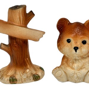 May include: A set of ceramic salt and pepper shakers. The salt shaker is shaped like a tree stump with a sign attached. The pepper shaker is shaped like a brown bear.