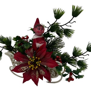 May include: A vintage Christmas decoration featuring a red and white elf riding in a white sleigh. The sleigh is surrounded by green pine branches, red holly berries, and a red poinsettia.