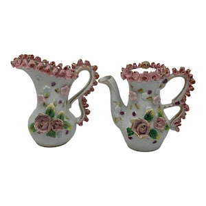 Może przedstawiać: Zestaw dwóch białych porcelanowych filiżanek do herbaty z różowymi i złotymi akcentami kwiatowymi. Filiżanki mają falbankę i uchwyt.