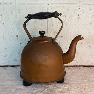 Peut inclure: Une bouilloire en cuivre vintage avec une poignée en bois noir et trois pieds noirs. La bouilloire a un corps arrondi et un bec verseur sur le côté.