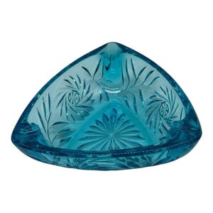 Puede incluir: Un cenicero triangular de vidrio turquesa con un detallado diseño floral y de espirales grabado. El cenicero tiene un diseño central en forma de sol y un borde curvo. El vidrio es de un azul vibrante y transparente.