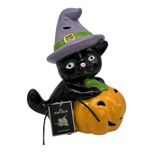 Puede incluir: Figura de gato negro con sombrero de bruja morado con banda verde y hebilla dorada, sosteniendo una calabaza naranja. La figura tiene una etiqueta que dice "GC HOME DECOR" y "pintado a mano".