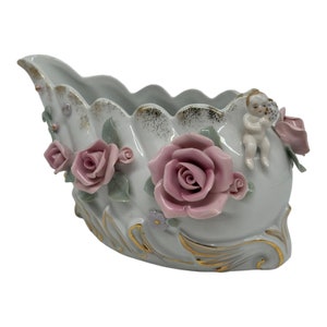 Könnte beinhalten: Eine weiße Porzellan Schale in Form einer Muschel mit rosa Rosen und einer Cherubfigur. Die Schale hat goldene Akzente und ist mit floralen Details verziert.