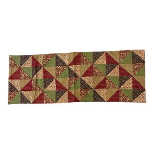 May include: A rectangular quilted table runner featuring a patchwork design of triangles in various colors and patterns. The colors include burgundy, green, tan, and brown, with floral and striped accents. The runner is approximately 60 inches long.